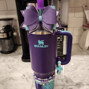 May include: A purple Stanley tumbler with a decorative bow, a jeweled top, and a beaded bracelet. The tumbler has a teal and silver bow, a cross charm, and a teal and purple base. The Stanley logo is visible on the front.