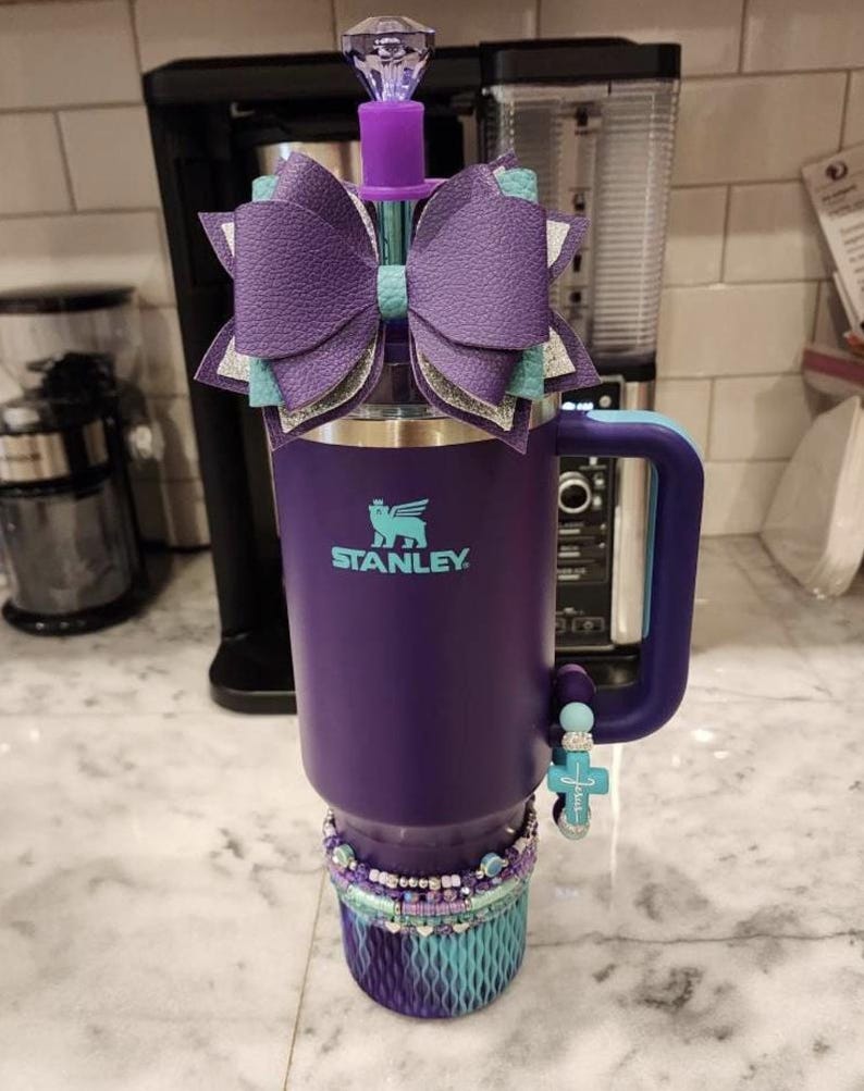 May include: A purple Stanley tumbler with a decorative bow, a jeweled top, and a beaded bracelet. The tumbler has a teal and silver bow, a cross charm, and a teal and purple base. The Stanley logo is visible on the front.