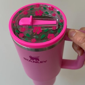 May include: Pink Stanley mug with a matching handle. The lid is clear with a pink floral design. The Stanley logo is visible on the side of the mug. The mug is designed to keep beverages hot or cold.