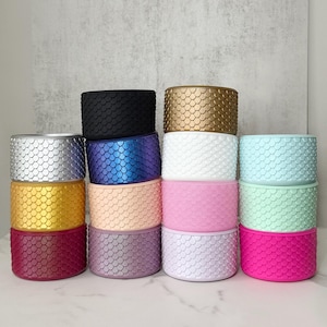 May include: A stack of cylindrical containers in various colors, including silver, gold, black, blue, white, pink, and green. Each container has a textured, scale-like pattern. The containers are stacked on a white surface against a neutral background.
