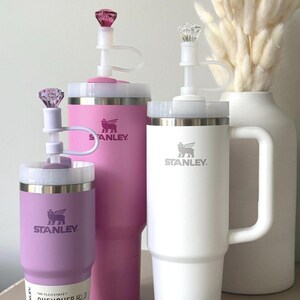 May include: Three Stanley Quencher H2.0 tumblers in varying sizes and colors: lavender, pink, and white. Each tumbler features a white lid, a clear straw, and a decorative gem on top. The Stanley logo is visible on each tumbler.