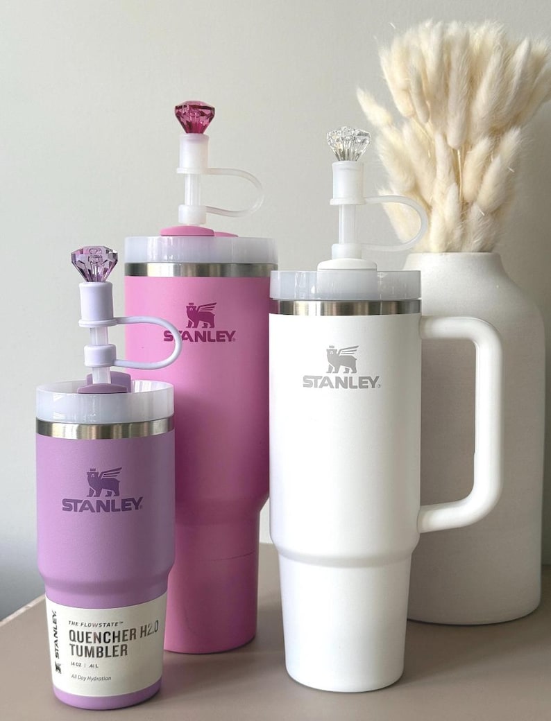 May include: Three Stanley Quencher H2.0 tumblers in varying sizes and colors: lavender, pink, and white. Each tumbler features a white lid, a clear straw, and a decorative gem on top. The Stanley logo is visible on each tumbler.