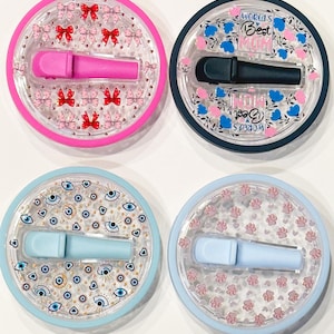 Pode incluir: Quatro recipientes redondos e transparentes com um aplicador cosm&eacute;tico no interior. Os recipientes t&ecirc;m diferentes desenhos: la&ccedil;os cor-de-rosa, o texto "World's Best Mum", olhos azuis e pegadas cor-de-rosa. Os aplicadores s&atilde;o cor-de-rosa, pretos, azul claro e azul claro.