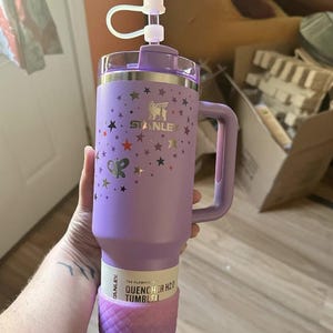 May include: A purple Stanley Quencher H2.0 tumbler with a handle, decorated with stars and a butterfly. The tumbler has a clear lid and a decorative top. The bottom of the tumbler is a textured, glittery pink. The text "STANLEY" is visible.