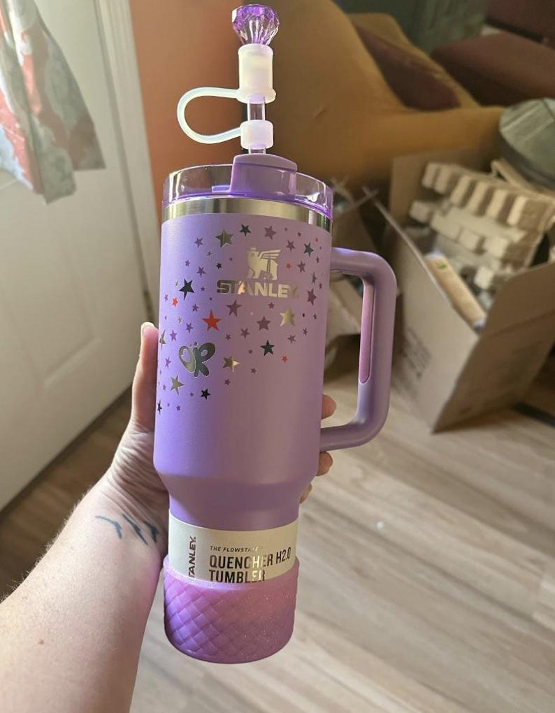 May include: A purple Stanley Quencher H2.0 tumbler with a handle, decorated with stars and a butterfly. The tumbler has a clear lid and a decorative top. The bottom of the tumbler is a textured, glittery pink. The text "STANLEY" is visible.