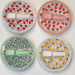 Pode incluir: Quatro recipientes redondos e transparentes com desenhos de frutas e flores. Cada um cont&eacute;m um aplicador labial em cores diferentes: branco, rosa, verde e branco. Os recipientes s&atilde;o decorados com morangos, p&ecirc;ssegos, abacates e lim&otilde;es.