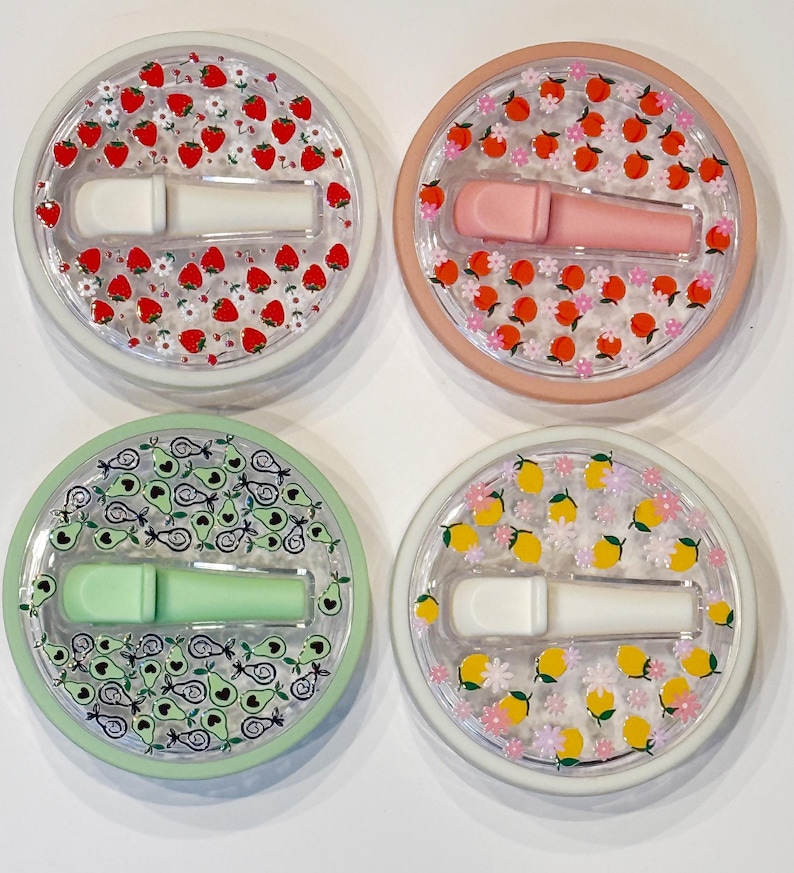 Pode incluir: Quatro recipientes redondos e transparentes com desenhos de frutas e flores. Cada um cont&eacute;m um aplicador labial em cores diferentes: branco, rosa, verde e branco. Os recipientes s&atilde;o decorados com morangos, p&ecirc;ssegos, abacates e lim&otilde;es.