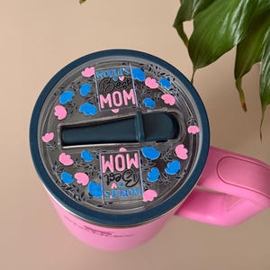 Pode incluir: Um copo t&eacute;rmico rosa com tampa e al&ccedil;a azul escuro. A tampa apresenta o texto "World's Best Mom" em uma fonte decorativa, rodeado por detalhes florais azuis e rosa. Para bebidas.