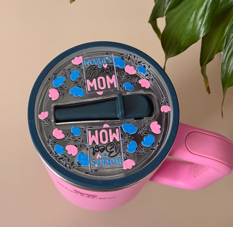 Pode incluir: Um copo t&eacute;rmico rosa com tampa e al&ccedil;a azul escuro. A tampa apresenta o texto "World's Best Mom" em uma fonte decorativa, rodeado por detalhes florais azuis e rosa. Para bebidas.
