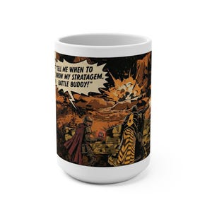 May include: White ceramic coffee mug featuring a comic book-style illustration of soldiers in a battlefield setting. The artwork includes a figure in a cape holding a glowing orb, with the text "TELL ME WHEN TO THROW MY STRATAGEM. BATTLE BUDDY!"