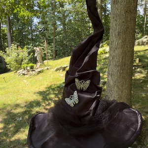 May include: A tall, pointed black hat with a wide brim, featuring a smooth satin-like finish and a gathered fabric texture. Two metallic gold-toned butterfly embellishments with intricate cut-out wing patterns are attached to the front cone.