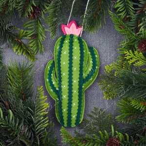 Może przedstawiać: Zielona ozdoba kaktusa z filcu z różową kwiatkiem z filcu na górze. Ozdoba jest zawieszona na sznurku i otoczona zielonymi gałązkami sosny.