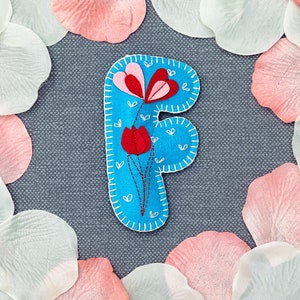 Könnte beinhalten: Ein blauer Filzbuchstabe F mit weißen Nähten, verziert mit einer roten und pinkfarbenen Filzblume mit weißen Stielen und weißen Herzen auf blauem Hintergrund.