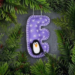 May include: A purple felt Christmas ornament in the shape of the letter "E" with white snowflake details and a penguin applique. The ornament is surrounded by evergreen branches.