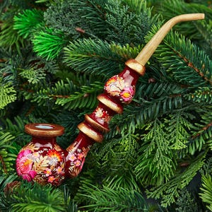 May include: A hand-painted tobacco pipe with a dark red bowl and stem, adorned with floral patterns in pink, yellow, and white. The pipe has a light brown, curved mouthpiece and gold accents. The pipe rests on a bed of green pine needles.