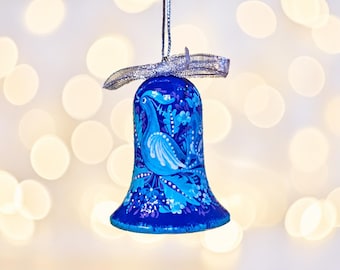 Puede incluir: Un adorno colgante en forma de campana con una base de color azul vibrante, decorado con motivos florales pintados a mano en azul claro y blanco, junto con un diseño de ave estilizada. Incluye una cinta metálica plateada para colgarlo.