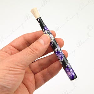 Puede incluir: Un bolígrafo negro con un patrón floral en morado y blanco, sostenido en una mano. El bolígrafo tiene una punta de color claro. El diseño floral es visible sobre un fondo blanco.