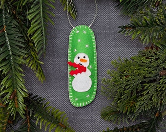 May include: A handmade Christmas ornament featuring a snowman design. The ornament is made of green felt with a white snowman wearing a red scarf. White stitching and small white stars decorate the green background. A silver string is attached for hanging.
