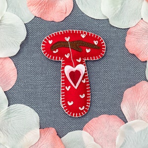 May include: A red felt mushroom with a brown felt branch and a white and red felt heart hanging from it. The mushroom is surrounded by pink and white rose petals.