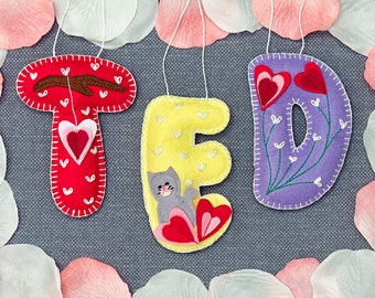 May include: Felt letter ornaments spelling the name "TED" in red, yellow, and purple. Each letter has a different design, including a heart, a cat, and flowers.