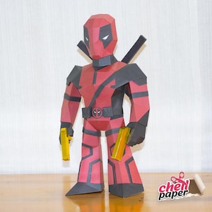 May include: A red and black papercraft model of Deadpool, a superhero character, holding two yellow guns. The model is made of folded paper and has a detailed design.