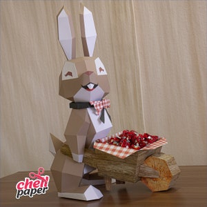May include: A brown papercraft bunny wearing a red and white bow tie pushes a wooden wheelbarrow filled with red and white sweets. The wheelbarrow has a red and white checked lining.