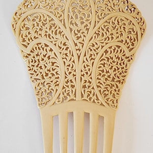 May include: Ivory-colored hair comb with intricate openwork design. The top section features a detailed, symmetrical pattern of swirling lines and floral motifs. The comb has five long, evenly spaced teeth.