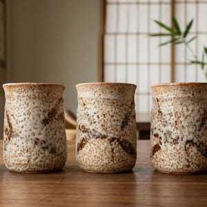 May include: Five ceramic tumblers with a speckled, textured surface in shades of beige and brown. Each tumbler has a cylindrical shape with a slightly flared rim. The design features a darker brown, leaf-like pattern. The tumblers are arranged on a wooden surface.