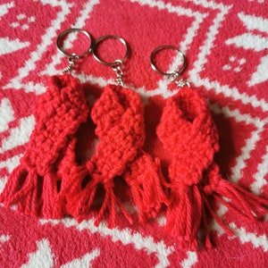 May include: Three handmade red crochet keyrings with silver rings. Each keyring features a rectangular design with a fringed bottom. The keyrings are displayed on a red and white patterned surface.