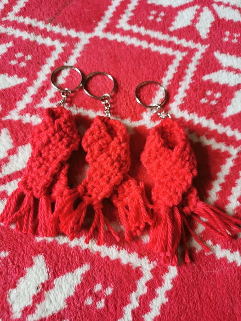 May include: Three handmade red crochet keyrings with silver rings. Each keyring features a rectangular design with a fringed bottom. The keyrings are displayed on a red and white patterned surface.