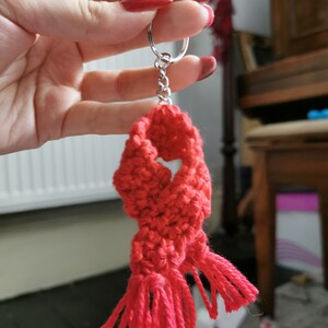 May include: A handmade red crochet scarf keyring with a silver ring. The scarf has a textured appearance with tassels at the bottom. The keyring is held up by a hand with red nail varnish.
