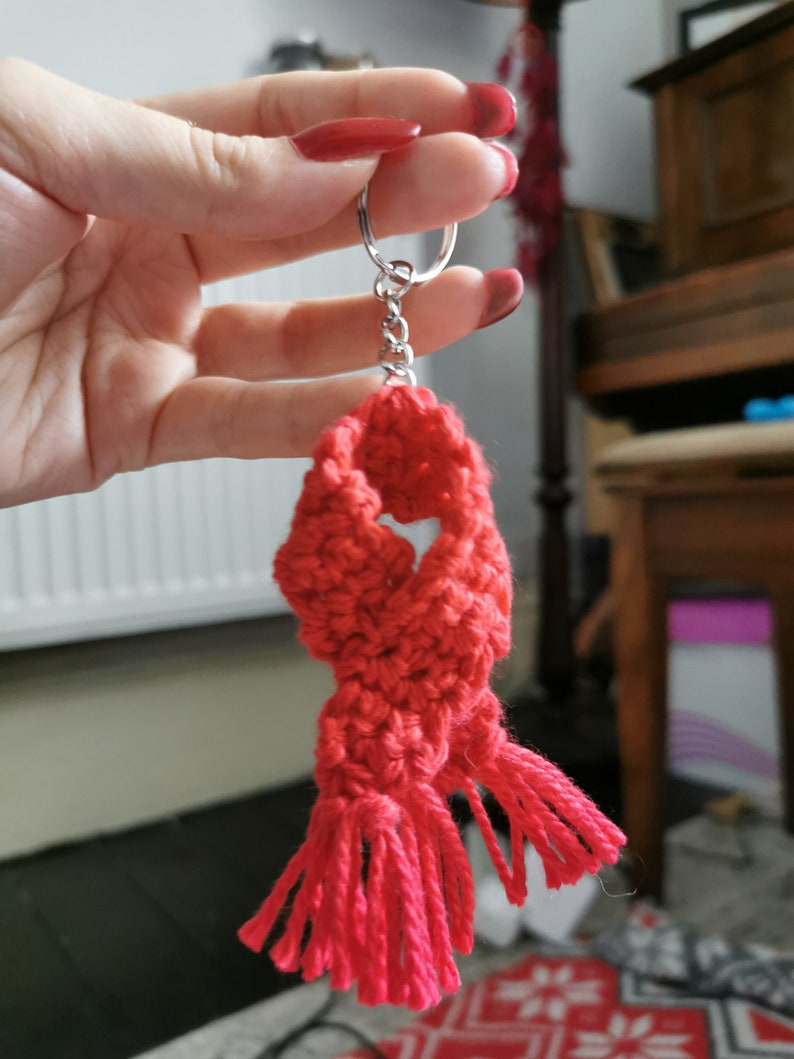 May include: A handmade red crochet scarf keyring with a silver ring. The scarf has a textured appearance with tassels at the bottom. The keyring is held up by a hand with red nail varnish.
