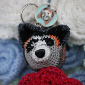 Könnte beinhalten: Ein gehäkelter Husky-Schlüsselanhänger in Schwarz, Weiß und Orange. Der Schlüsselanhänger ist an einem silbernen Schlüsselring mit einem runden, blau-weißen Emaille-Anhänger befestigt.