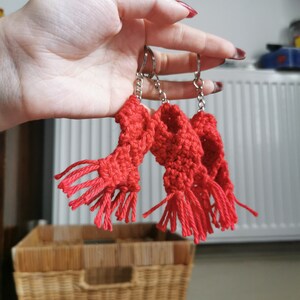 May include: Three red crocheted scarf keyrings with silver keyrings and chains. The scarves have fringed ends. The keyrings are held in a hand against a background of a white radiator, a wicker basket, and a wall.