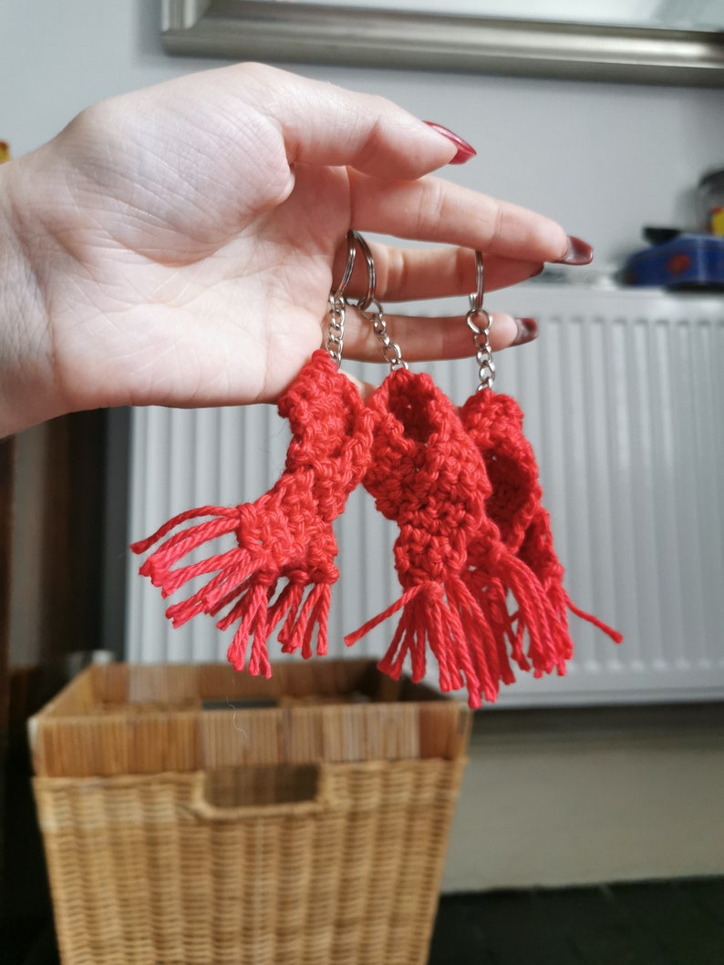 May include: Three red crocheted scarf keyrings with silver keyrings and chains. The scarves have fringed ends. The keyrings are held in a hand against a background of a white radiator, a wicker basket, and a wall.