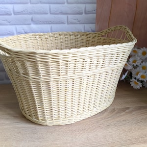 May include: A large, oval-shaped wicker basket with handles. The basket is made of natural woven wicker and has a light cream color.