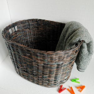 Puede incluir: Una cesta de lavandería de mimbre grande, de forma ovalada, con un acabado marrón oscuro y verde. La cesta está hecha de mimbre tejido y tiene un asa a cada lado. Una toalla gris está colgada sobre el borde de la cesta.