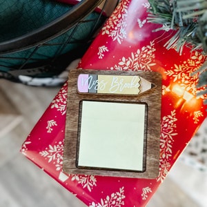 Puede incluir: Un letrero de madera con forma de l&aacute;piz con una nota adhesiva en blanco y el texto "Miss Brooke" en la parte superior. El letrero est&aacute; sobre papel de regalo rojo y blanco con un patr&oacute;n floral.