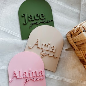 May include: Three arched name plaques with personalized names. The plaques are green, beige, and pink. The names are "Jace Parker", "Aria Lynn", and "Alaina Grace".