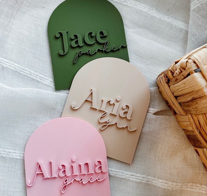 May include: Three arched name plaques with personalized names. The plaques are green, beige, and pink. The names are "Jace Parker", "Aria Lynn", and "Alaina Grace".