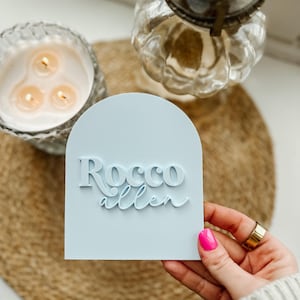May include: A light blue arch-shaped sign with the text "Rocco Allen" in white raised lettering.