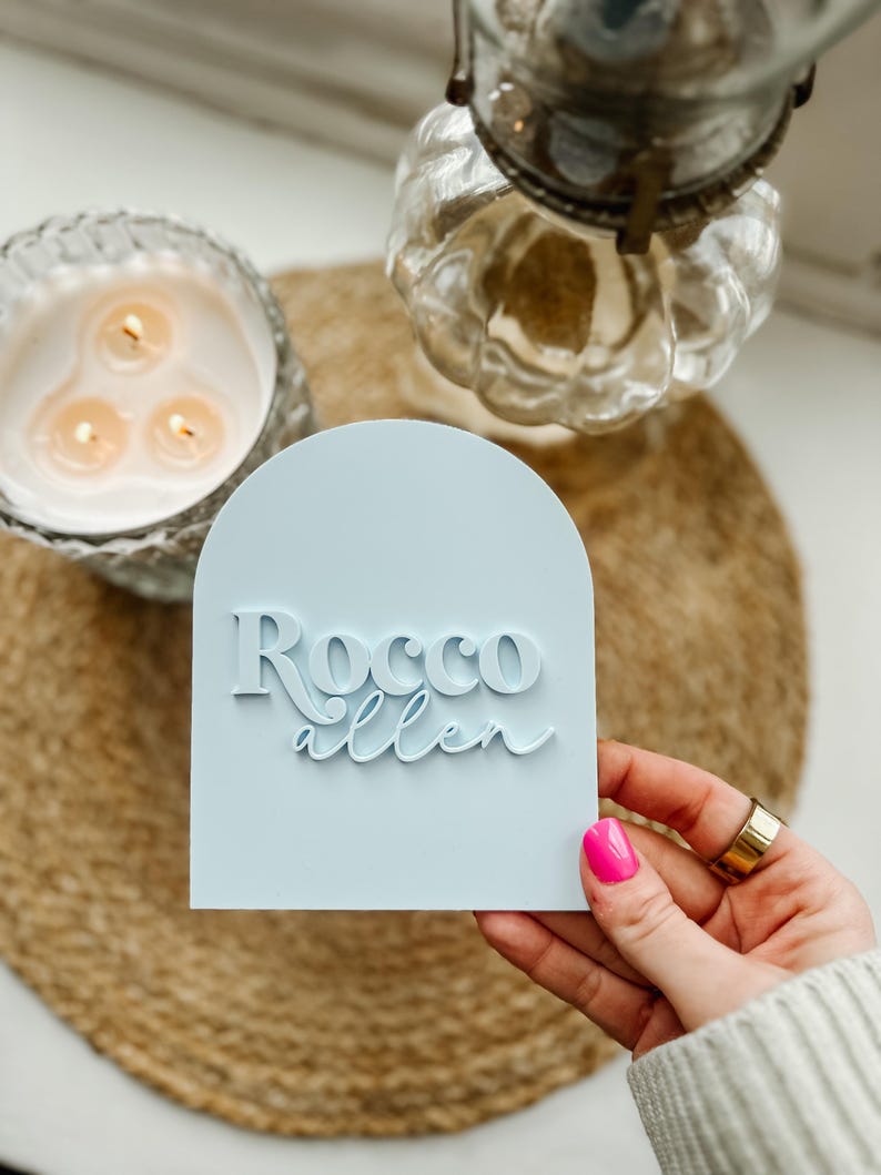 May include: A light blue arch-shaped sign with the text "Rocco Allen" in white raised lettering.