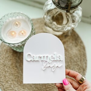 May include: A white card with a pink background and the names "Carmela Gigi" written in white lettering.