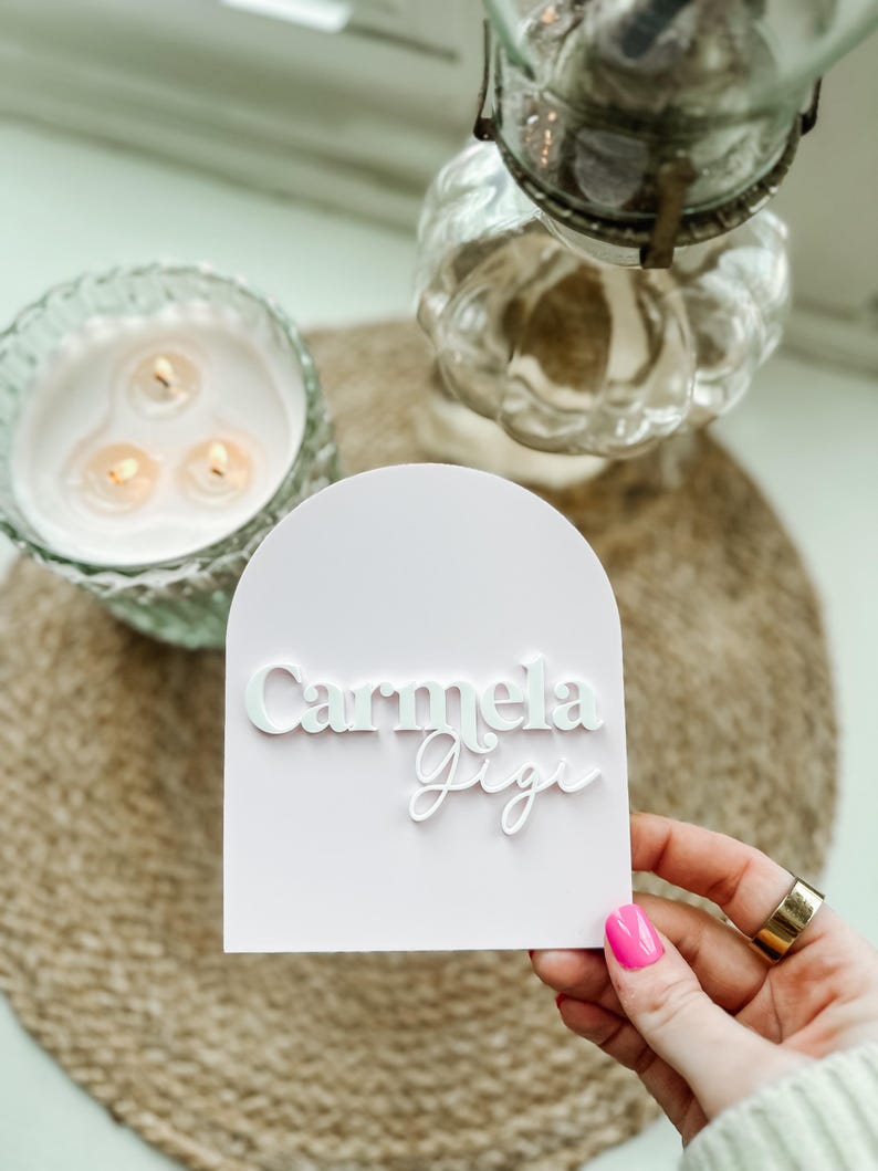 May include: A white card with a pink background and the names "Carmela Gigi" written in white lettering.