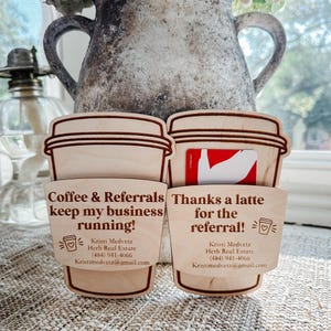 Può includere: Due porta biglietti da visita in legno a forma di tazza di caffè. Uno dice "Coffee & Referrals keep my business running!" e l'altro "Thanks a latte for the referral!". Entrambi hanno le informazioni di contatto di Kristi Medvetz, Herb Real Estate.