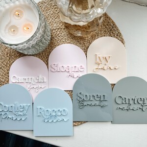 May include: Seven pastel-colored arch-shaped name plaques with names written in white and black lettering. The names are: Carmela, Sloane, Ivy Jane, Soren Brooks, Carter Robert, Conley Pratt, and Rocco Allen.