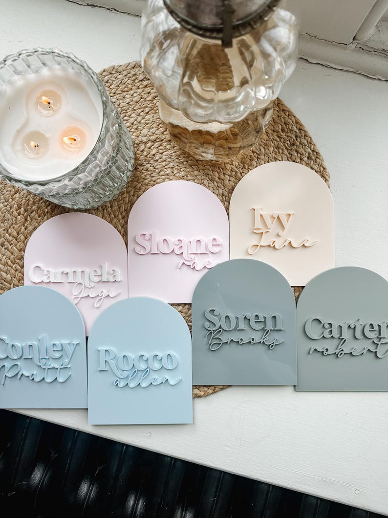 May include: Seven pastel-colored arch-shaped name plaques with names written in white and black lettering. The names are: Carmela, Sloane, Ivy Jane, Soren Brooks, Carter Robert, Conley Pratt, and Rocco Allen.
