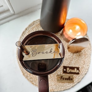May include: A brown travel mug with a gold lid and a gold name tag that says "Brooke". The mug is sitting on a woven mat with two other gold name tags that say "Brooke".