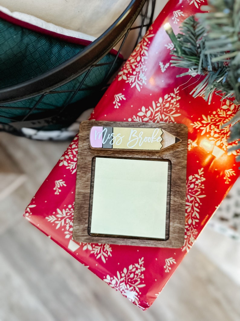 Puede incluir: Un letrero de madera con forma de l&aacute;piz con una nota adhesiva en blanco y el texto "Miss Brooke" en la parte superior. El letrero est&aacute; sobre papel de regalo rojo y blanco con un patr&oacute;n floral.