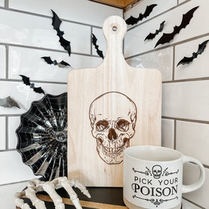 May include: A wooden cutting board with a skull design and the text "Pick Your Poison" on a white mug. The cutting board and mug are on a black and brown round tray with a skeleton hand and a black spiderweb dish.